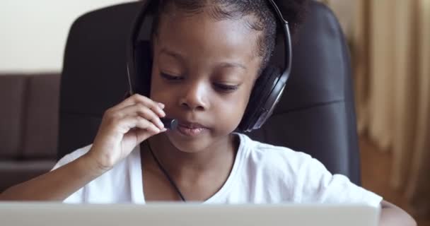 Little African girl plays at laptop child pretends call manager, schoolgirl daughter wears headphones on her head and listens to music while sitting at home table, baby passionate about computer games - Footage, Video