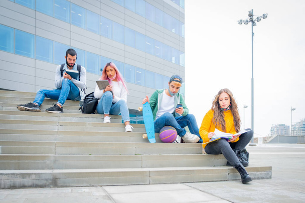 Νέοι που σπουδάζουν στο πανεπιστήμιο κατά τη διάρκεια της πανδημίας του κορωναϊού. Τέσσερις πολυετείς φοιτητές πανεπιστημίου με προστατευτικές μάσκες κάθονται έξω στις σκάλες κρατώντας την κοινωνική απόσταση - Φωτογραφία, εικόνα