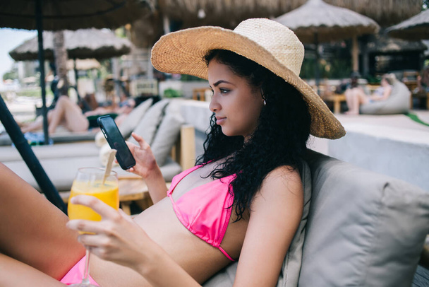 Seitenansicht der schönen jungen ethnischen Frau im trendigen Bikini und Strohhut mit dem Handy beim Liegen auf der Sonnenliege und Blick weg auf den Badeort - Foto, Bild