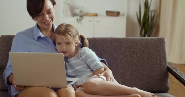Caucasian mom with cute daughter sitting on comfortable sofa in living room at home hugging watching cartoons together in laptop online laughing spend time together, happy family motherhood concept - Footage, Video