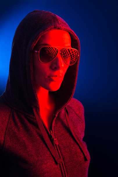 Retrato de una joven con capucha y gafas de sol, iluminadas con una luz roja, sobre un fondo azul oscuro. - Foto, Imagen