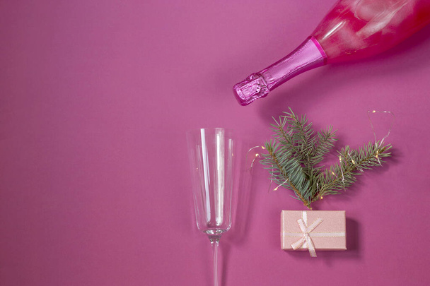 Festive New Year concept. A bottle of pink champagne and a glass for it on a pink background. Christmas tree, garland and gift box. Copy space. Postcard top view. flat lay - Photo, Image