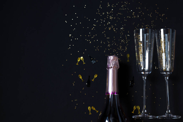 A bottle of champagne and two glasses, golden confetti on a festive black background. Festive concept. Copy space. - Photo, Image