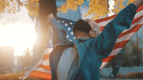 Młoda kobieta z maską na twarzy dumnie trzymająca amerykańską flagę na świeżym powietrzu.USA koncepcja wyborów prezydenckich.  - Zdjęcie, obraz