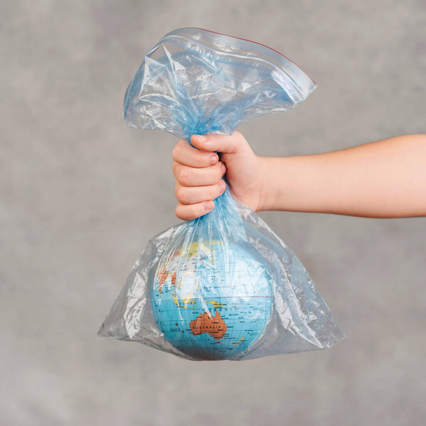 dětská ruka drží zeměkouli v průhledném plastovém sáčku přes šedé pozadí - Fotografie, Obrázek