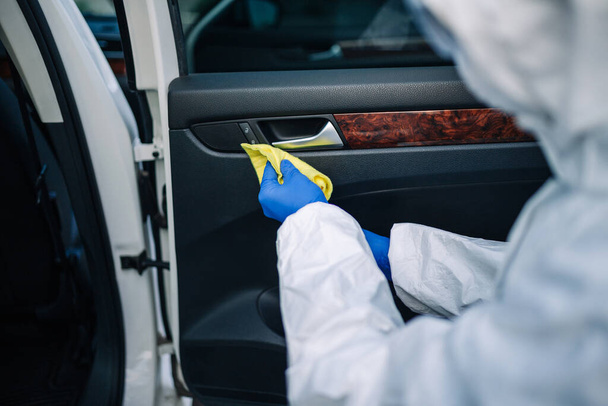 Υγιεινής εργαζόμενος υπηρεσία καθαρίζει το εσωτερικό του αυτοκινήτου με ένα κίτρινο χαλί. Ένας άντρας με προστατευτική στολή, μάσκα και γάντια απολυμαίνει τις πόρτες του οχήματος. Coronavirus covid έννοια της πρόληψης εξάπλωσης - Φωτογραφία, εικόνα
