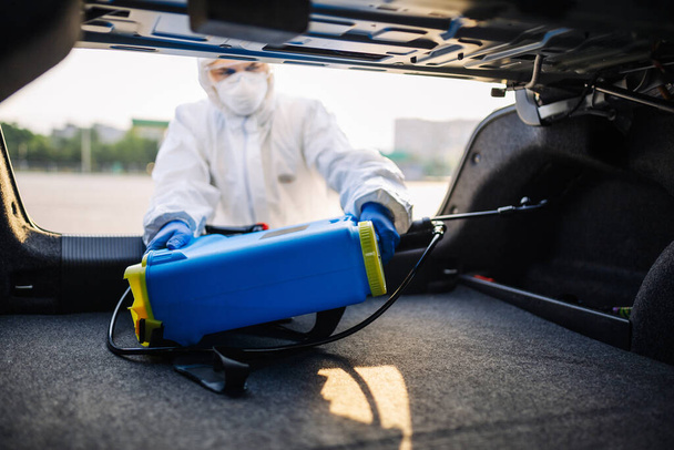 Disinfection service worker puts a spray equipment into a vehicle's trunk. A man wearing protective suit, mask and gloves takes out a sanitary spray from a car. Cleaning and covid-19 prevention - Foto, afbeelding