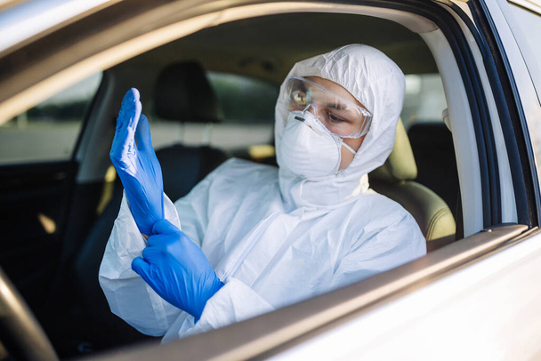 Desinfektionsarbeiter zogen Handschuhe an, um die Ausbreitung von Kolibakterien zu stoppen. Ein Mann in Schutzanzug, Maske und blauen Handschuhen in einem Auto. Desinfektion und Gesundheitskonzept - Foto, Bild