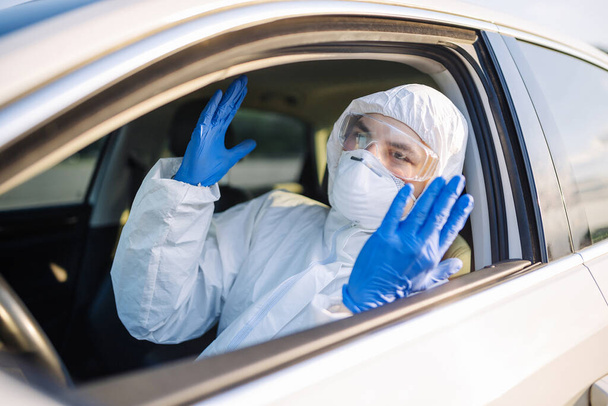Ο απολυμαντής εξοργίστηκε λόγω της παγκόσμιας επιδημίας κοπριάς. Ένας άντρας που φοράει προστατευτική στολή, μάσκα και μπλε γάντια σε ένα αυτοκίνητο. Απολύμανση και έννοια της υγείας - Φωτογραφία, εικόνα