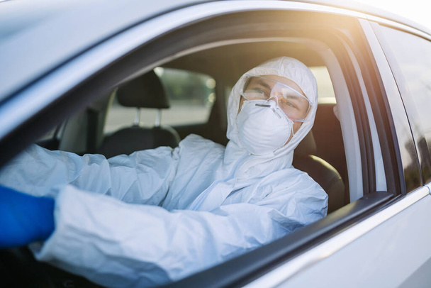 Desinfectiewerker rijdt in een auto tijdens wereldwijde covid-19 pandemische uitbraak quarantaine. Een man met een beschermend pak, masker en blauwe handschoenen in een auto. Desinfectie- en gezondheidsconcept - Foto, afbeelding