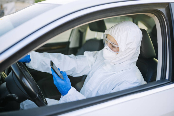 Desinfektionsarbeiter benutzt sein Mobiltelefon während einer Autofahrt während des Ausbruchs der Covid-Pandemie. Ein Mann in Schutzanzug, Maske und blauen Handschuhen in einem Auto. Desinfektion und Gesundheitskonzept - Foto, Bild