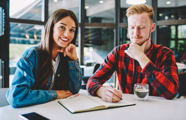 Portrait of happy Caucasian hipster girl smiling at camera during private lesson with intelligent red hair man writing plan organisation in education textbook, couple learning together at coffee shop - Foto, Bild