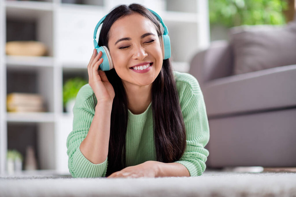 Close up photo of charming chinese girl listen music use blue wireless headset enjoy sound lie floor carpet in house indoors - Foto, afbeelding