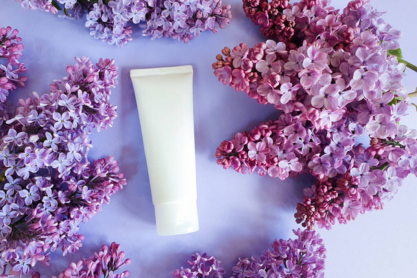 Top view of mockup of white squeeze bottle plastic tube for branding of medicine or cosmetics - cream, gel, skin care. Cosmetic bottle container on and lilac flowers on light purple table. - Photo, Image