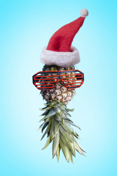 Санта-Ананас. Поп-арт в летней концепции минимален. Забавный ананас в солнечных очках и шляпе Санта Клауса на ярком пастельном фоне. Холодный праздничный креативный дизайн. - Фото, изображение