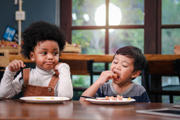 Cute African American boy with curly and adorable Asian kid eating meal at the table indoor, happy children having food in a restaurant, enjoy their meal together.  - Foto, afbeelding