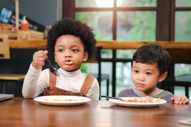 Cute Afroamerykanin chłopiec z kręcone i urocze azjatyckie dziecko jedzenie posiłek przy stole kryty, szczęśliwe dzieci posiadające jedzenie w restauracji, cieszyć się posiłkiem razem.  - Zdjęcie, obraz