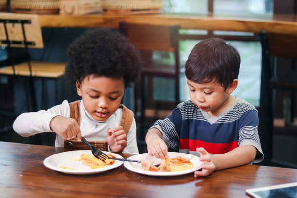 Cute African American boy with curly and adorable Asian kid eating meal at the table indoor, happy children having food in a restaurant, enjoy their meal together.  - Photo, Image