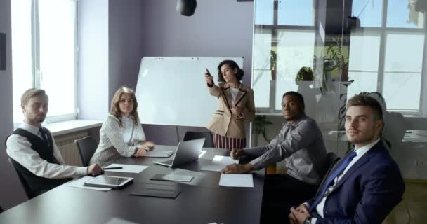 Business woman presenting project to colleagues multinational group showing information screen. Female leader coach speaks at office conference points to digital board switches slide by remote control - Footage, Video