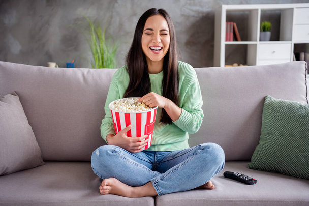 Full body photo of candid pretty korean girl sit divan legs bare foot crossed watch funny series laughing eat pop corn box in house indoors - Photo, image