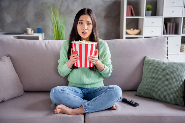 Full length photo of worried chinese girl sit divan legs crossed scared horror series hold big pop corn box in house indoors - Фото, изображение