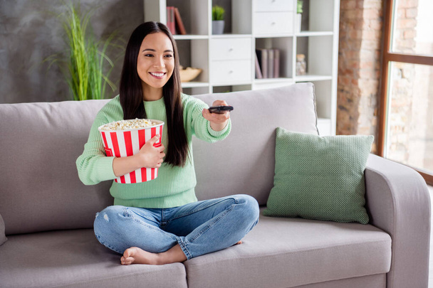 Full length photo of positive cheerful korean girl sit divan legs crossed watch film switch channel remote control hold big pop corn box in house indoors - Fotó, kép