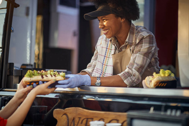  empleada afro-americana en servicio de comida rápida dando bocadillos ordenados a un cliente con una sonrisa - Foto, Imagen