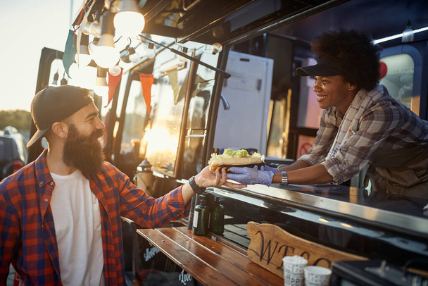 afro-american female employee gives with care and smile a sandwich to young hipster male customer - 写真・画像