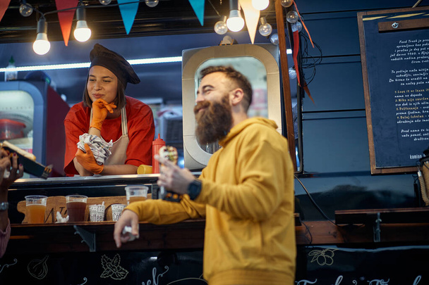 young beardy hipster eating sandwich in front of fast food service, socializing with female employee - Foto, Bild