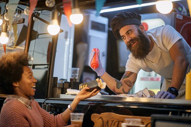 young beardy caucasian employee in fast food adding a ketchup in a sandwich to a female afro-american customer, joking, amusing - Foto, afbeelding