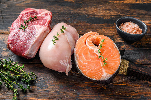 Different types of raw meat steaks Beef striploin, salmon and chicken breast. Dark wooden background. Top view. - Photo, Image