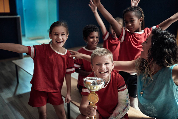 Kids team and female coach excited at the locker room about won trophy. Children team sport - Fotoğraf, Görsel