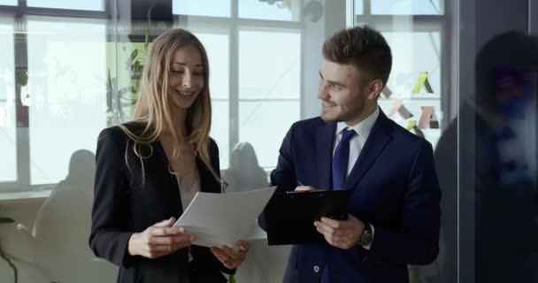 Woman in formal suit and business man two people colleagues standing in corridor of office looking at papers documents talking laughing informal communication in company, colleagues discussing project - Footage, Video