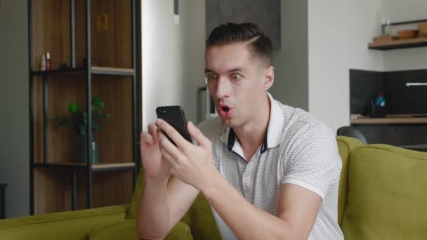 Surprised young man winner hold smartphone read good news amazed by mobile online bet bid game win at home. Happy male look at cell phone screen overjoyed by victory success while sitting on sofa. - Footage, Video