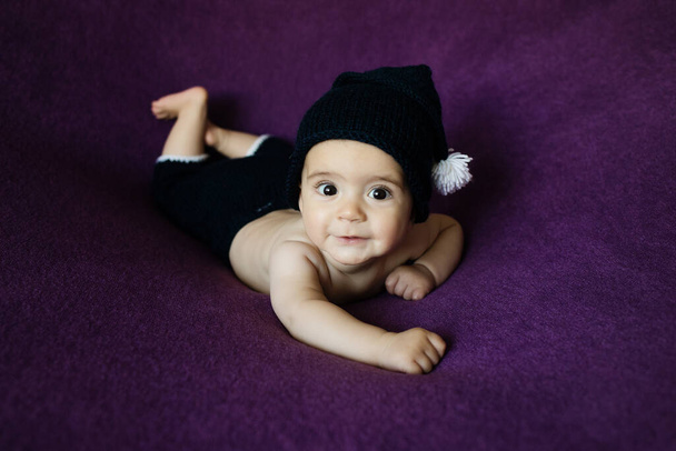 Симпатичный новорожденный в тёмно-синей шляпе. Счастливого малыша на фиолетовом фоне. Портрет новорожденного крупным планом. Шаблон упаковки детских товаров. Детская. Медицинская и здоровая концепция. Рождество. Новый год - Фото, изображение