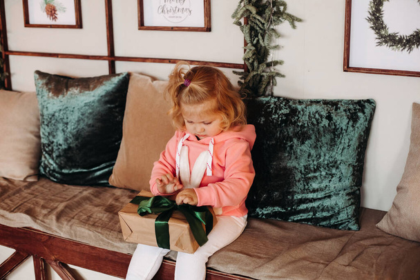 Petite jolie fille Sith cheveux ondulés assis sur le canapé ouvre une boîte avec un cadeau - Photo, image
