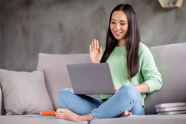 Full length photo of positive girl korean social media blogger sit couch legs crossed use laptop have online live broadcast wave hand in house indoors - Photo, image