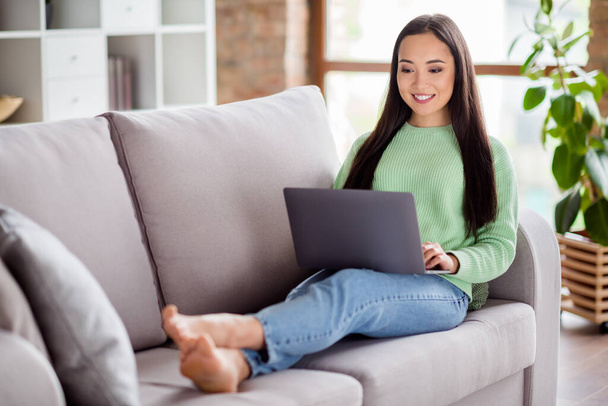Full size photo of positive korean girl university student study remote work laptop prepare project wear green sweater denim jeans bare foots lie divan in house indoors - Photo, Image