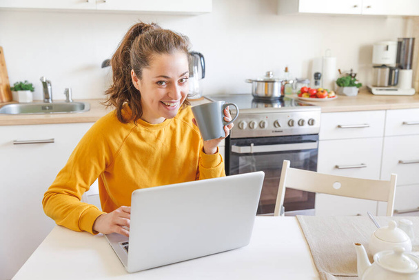 Oficina Móvil en casa. Mujer joven sentada en la cocina en casa trabajando con un ordenador portátil. Chica de estilo de vida estudiando o trabajando en interiores. Concepto de cuarentena empresarial independiente. - Foto, imagen