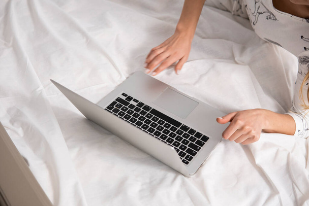 Top view of a woman sitting on a bed , using laptop. Working at home concept. Girl is working on her laptop in bed with white linens. Relaxing working process. Stay at home. - Foto, afbeelding