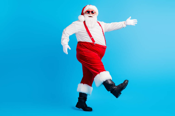Full size profile side photo of funky santa claus go copyspace x-mas christmas holly newyear wear suspenders overalls sunglass boots cap isolated over blue color background - Fotografie, Obrázek
