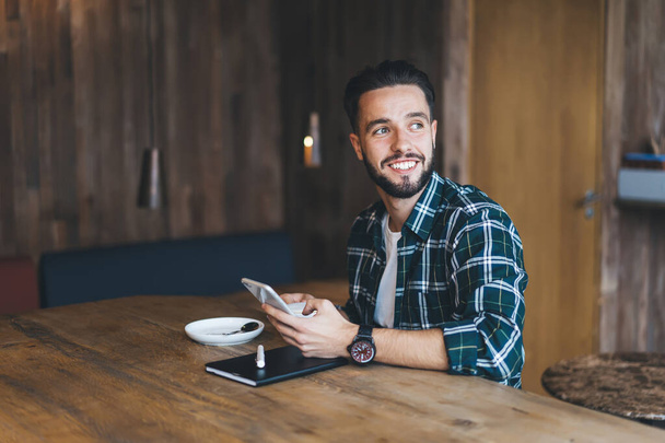 Веселый мужчина с бородой и усами в повседневной одежде, работающий со смартфоном и записками, наслаждаясь кофе в уютном кафе - Фото, изображение
