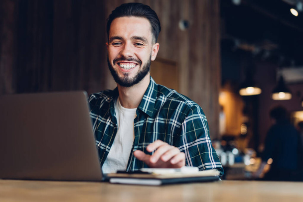Happy bearded male freelancer in checkered shirt chatting on laptop with colleague while sitting at table with notebook in loft cafeteria in evening and smiling looking at screen - Foto, imagen