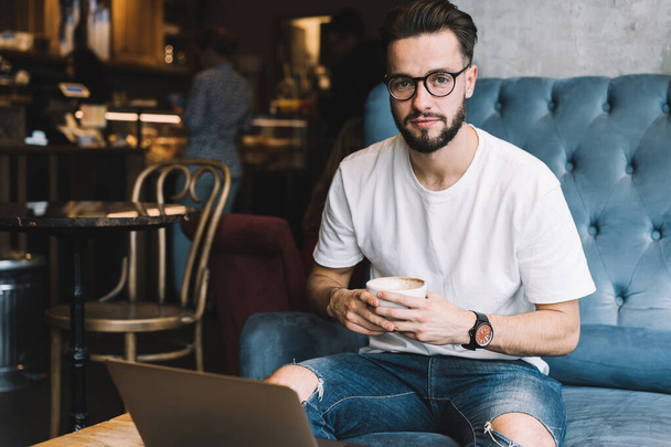 Youthful bearded male in casual wear and glasses drinking coffee and using laptop while sitting at table in cafe and looking at camera - Photo, image
