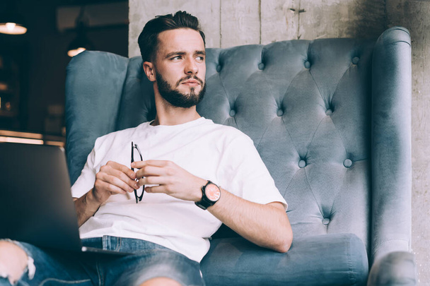 Pensive bearded male freelance worker in t shirt and jeans sitting on armchair in cafe with eyeglasses in hand and looking away - Fotoğraf, Görsel