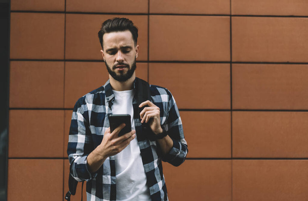 Puzzled male hipster with backpack wearing white t shirt and checkered shirt browsing mobile phone while standing in front of wall - Фото, изображение