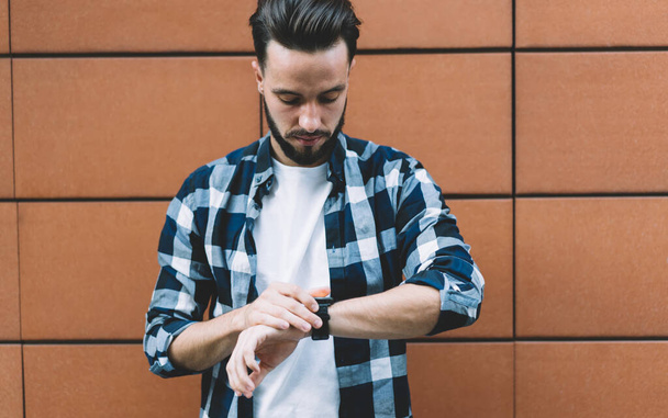 Focused bearded young man in style standing near building wall and looking down at wristwatch checking time outside in daytime - Foto, Bild