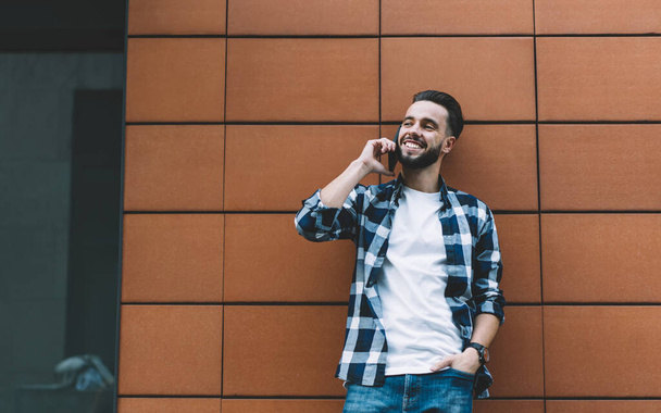 From below of smiling bearded young male in casual wear talking on cellphone while standing next to contemporary urban building and looking away on city street - Foto, afbeelding