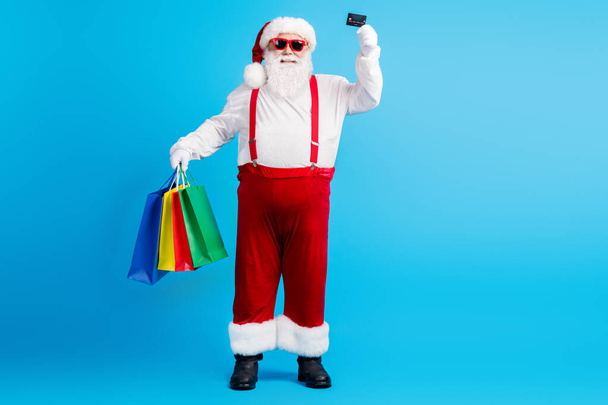 Full size photo of stylish white grey hair bearded santa claus hold credit card buy x-mas christmas bargain boutique bags wear red suspenders overall isolated blue color background - Valokuva, kuva