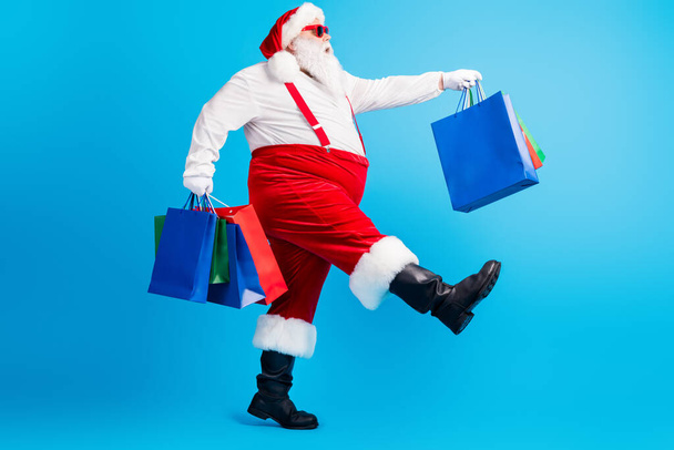Full body profile side photo of crazy white grey hair bearded fat santa claus go shopping hold x-mas christmas bags wear red suspenders overalls isolated over blue color background - Foto, afbeelding